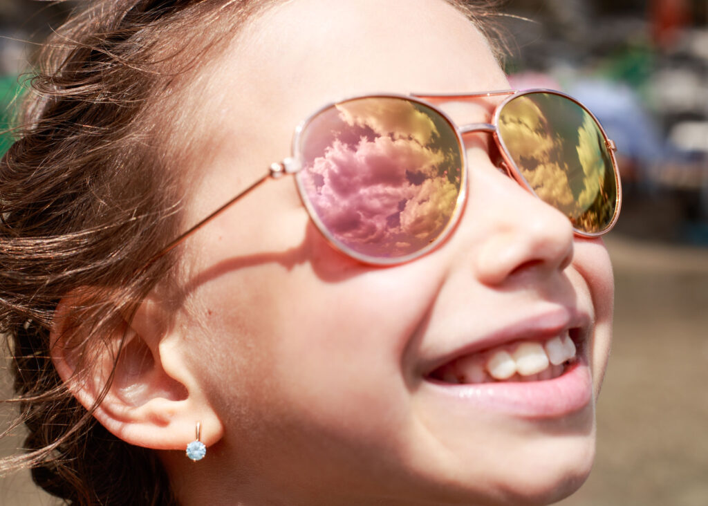 Twarz dziewczynki. W okularach przeciwsłonecznych odbijają się chmury